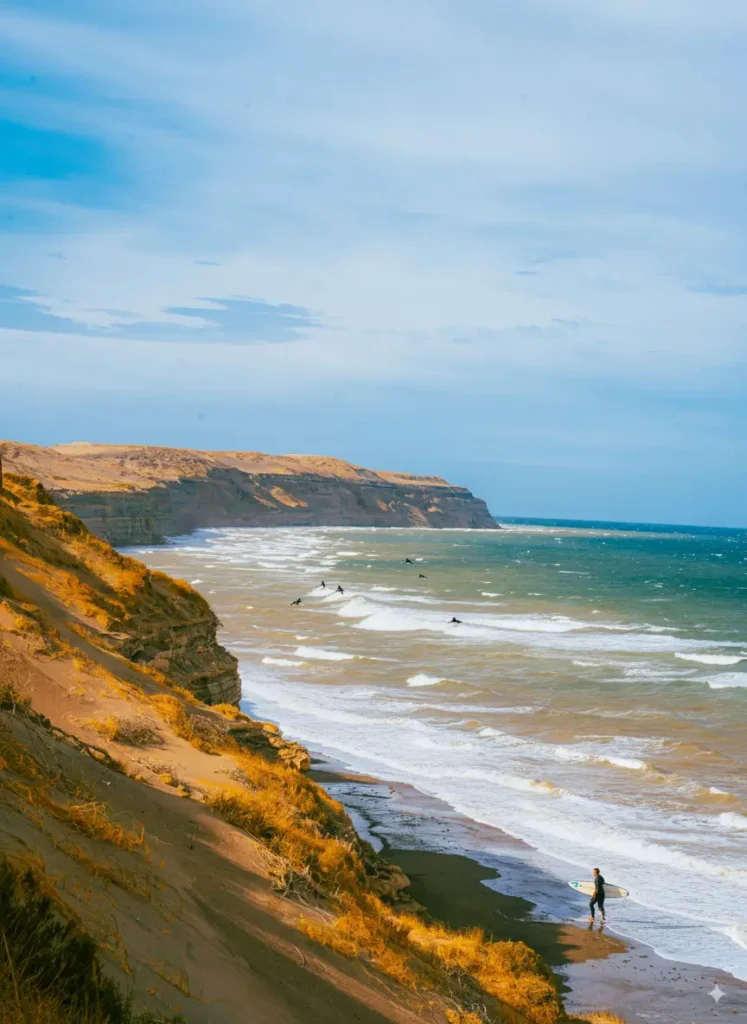 Surfer walking along the golden coastline of Lobitos Beach in Peru with waves, cliffs, and surfers in the water – a top surfing spot on Peru Beaches.